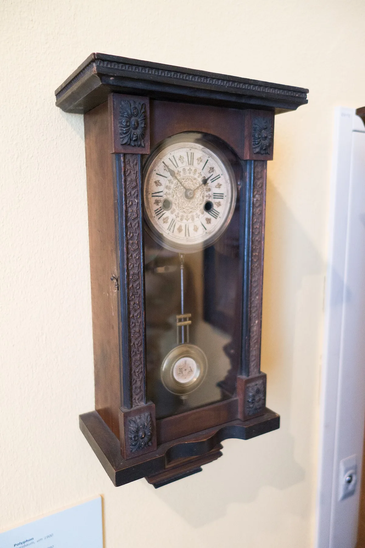 Antike Pendel-Wanduhr aus dunklem Holz mit vergilbtem Zifferblatt an einer hellen Wand